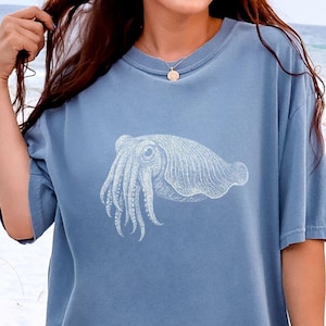 May include: A blue jean colored t-shirt featuring a white line art illustration of a cuttlefish centered on the chest. The garment has a crew neckline, short sleeves, and a relaxed fit. The text Blue Jean is printed in the bottom right corner.
