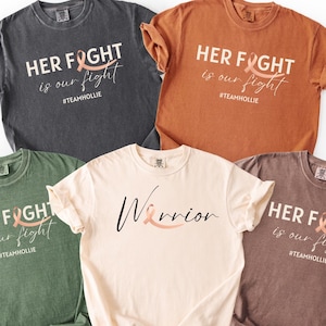 May include: Five different colored t-shirts are displayed, each with a unique design. The shirts are in shades of gray, orange, green, beige, and brown. The text on the shirts reads "HER FIGHT is our fight #teamhollie" and "Warrior".