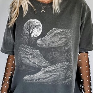 May include: A charcoal gray short-sleeve t-shirt featuring a white graphic print of three alligators in a swamp under a full moon. The shirt is layered over black sheer long sleeves adorned with small white pearls. The word Pepper is printed.