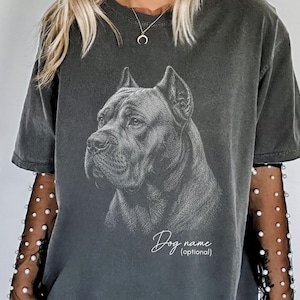 Pode incluir: Uma t-shirt cinzento-carvão de manga curta com uma ilustração de linhas brancas de uma cabeça de cão Cane Corso. A t-shirt é usada sobre mangas compridas pretas transparentes adornadas com pequenas pérolas brancas. O texto "Dog name (optional)" está impresso por baixo.
