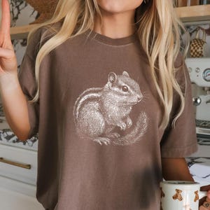 May include: Espresso-colored t-shirt featuring a detailed white illustration of a chipmunk. The shirt is paired with a mug adorned with teddy bear graphics. The overall aesthetic is casual and comfortable.