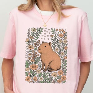 Puede incluir: Camiseta rosa claro con una ilustraci&oacute;n de carpincho marr&oacute;n rodeada de flores y estrellas. El carpincho est&aacute; sentado erguido dentro de un marco floral rectangular. La camiseta es de un material suave y c&oacute;modo.