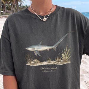 May include: A charcoal gray short-sleeve t-shirt featuring a detailed illustration of a thresher shark swimming above a sandy seabed with seaweed. The text "Thresher shark (Alopias vulpinus)" is printed in a white serif font below the shark graphic.
