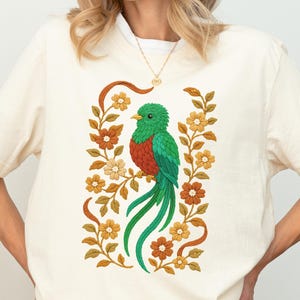 Op de afbeelding: Crèmekleurig T-shirt met een kleurrijke illustratie van een quetzalvogel. De vogel heeft groene, rode en blauwe veren en zit op een tak, omringd door oranje en crèmekleurige bloemen. Het ontwerp wordt omlijst door een bloemenrand.