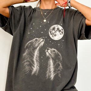 Può includere: Una t-shirt a maniche corte color grigio antracite con una stampa grafica bianca di due cani che guardano verso una luna piena circondata da stelle. La maglia presenta un girocollo e una vestibilità ampia, con la parola Pepper in un cerchio.