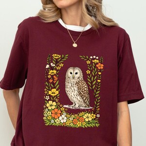 May include: Burgundy t-shirt with a white collar featuring a detailed illustration of an owl surrounded by colorful flowers. The owl is depicted in shades of brown and white, set against a floral border with yellow, orange, and white blossoms.