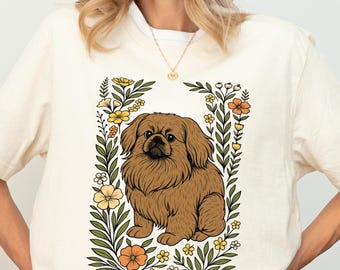 May include: Cream-colored t-shirt featuring a brown Pekingese dog illustration within a floral frame. The dog has fluffy fur and a friendly expression. The flowers are yellow and orange, with green leaves. The shirt has short sleeves.
