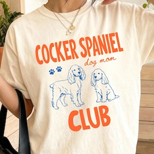 May include: Ivory t-shirt with the text "COCKER SPANIEL dog mom CLUB" in orange and blue. The shirt features illustrations of two Cocker Spaniel dogs and paw prints. The t-shirt is a casual style.