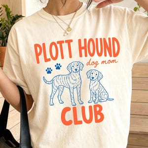 May include: Ivory t-shirt with the text "PLOTT HOUND dog mom CLUB" in orange and blue. The shirt features a graphic of two Plott Hound dogs. The t-shirt is a comfortable, casual style.