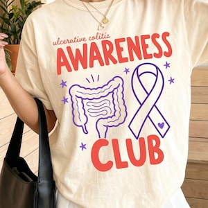 May include: Ivory-colored t-shirt with the words "ulcerative colitis AWARENESS CLUB" in red and purple. The shirt features a graphic of intestines and a purple awareness ribbon. The model is wearing a white pleated skirt.