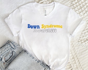 Puede incluir: Camiseta blanca con las palabras "Down Syndrome Awareness" en letras azules y amarillas. Unos vaqueros de mezclilla de lavado claro se colocan junto a la camiseta doblada. El texto de la camiseta está en una fuente retro.
