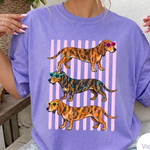 May include: A light purple t-shirt featuring a graphic of three dachshund dogs standing in a row. Each dog has a unique coat pattern and wears colourful sunglasses, including pink, teal, and yellow frames, set against a background of vertical pink stripes.