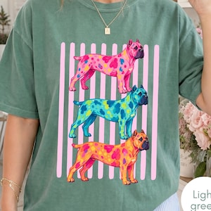 May include: A sage green short-sleeve t-shirt featuring a graphic print of three dogs stacked vertically. The dogs are rendered in vibrant, multi-coloured patterns against a background of vertical light pink stripes on the front of the shirt.