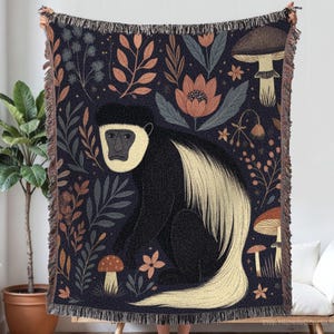 Może przedstawiać: Tkany koc z ilustracją czarno-białej małpy otoczonej kwiatami i grzybami na ciemnoniebieskim tle. Koc ma frędzelkową krawędź i jest trzymany przez osobę.