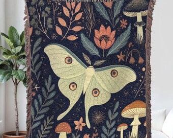 May include: A dark blue woven throw blanket featuring a large pale green moth with brown and yellow details. The blanket has a botanical design with mushrooms, flowers, and leaves in shades of orange, green, and brown. The edges have a fringed trim.