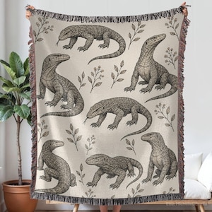 Può includere: Una coperta tessuta color beige chiaro con un motivo ripetuto di illustrazioni a linee nere raffiguranti draghi di Komodo e piccoli rametti con foglie. La coperta presenta una superficie testurizzata e bordi decorativi con frange.