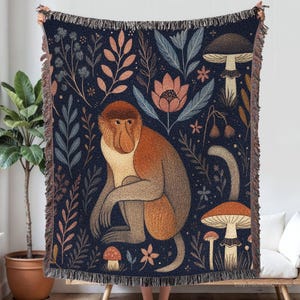 May include: A woven throw blanket featuring a whimsical design of a proboscis monkey, mushrooms, and floral elements in shades of brown, orange, and blue. The blanket has a fringed edge and is held up by a person.