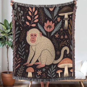 Könnte beinhalten: Eine gewebte Decke mit einem beigen Affen-Design, umgeben von Pilzen, Blumen und Blättern in Braun-, Rosa- und Blautönen auf dunklem Hintergrund. Die Decke hat einen Fransenrand und wird von einer Person gehalten.
