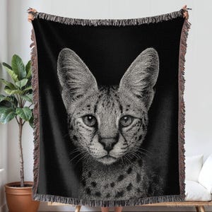 Puede incluir: Una manta tejida en blanco y negro que presenta un retrato detallado de un serval con orejas grandes y pelaje manchado. La manta tiene flecos texturizados a lo largo de los bordes superior e inferior, mostrando un diseño monocromático.