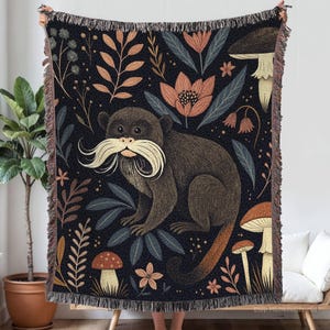 Può includere: Una coperta scura tessuta con un'illustrazione stravagante di una scimmia con lunghi baffi bianchi. La scimmia è circondata da fiori, funghi e fogliame nei toni dell'arancione, del verde e del marrone. La coperta ha un bordo sfrangiato.
