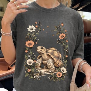 Op de afbeelding: Een antracietgrijs T-shirt met korte mouwen en een grafische print van een bruine schildpad in het midden van een kader van oranje en witte bloemen en kleine gouden sterren. Het T-shirt heeft een ronde hals en een relaxte pasvorm.