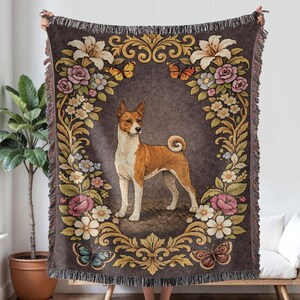May include: A woven throw blanket featuring a Basenji dog illustration within a floral and butterfly border. The blanket has a dark brown background with cream, pink, and gold accents. The edges are fringed.