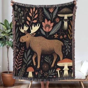 Puede incluir: Una manta tejida con un diseño de un alce marrón rodeado de plantas y setas estilizadas sobre un fondo negro. La manta tiene un borde con flecos. La paleta de colores incluye tonos de marrón, naranja y verde. Una persona sostiene la manta.