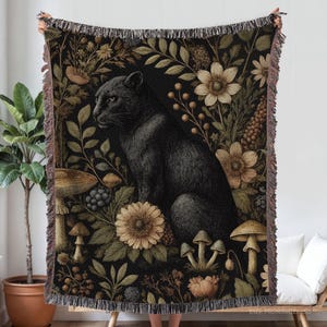 May include: A black panther sits amidst a floral tapestry of mushrooms, flowers, and foliage. The throw blanket features a dark background with a detailed botanical design and fringed edges. The overall color palette includes shades of black, brown, and beige.