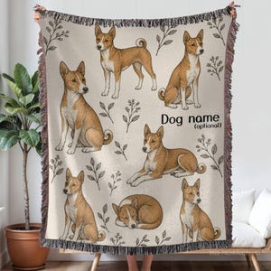 May include: A woven throw blanket featuring a repeating pattern of tan and white Basenji dogs in various poses, including sitting, lying down, and sleeping, interspersed with small leaf sprigs. The text "Dog name (optional)" is printed in black.