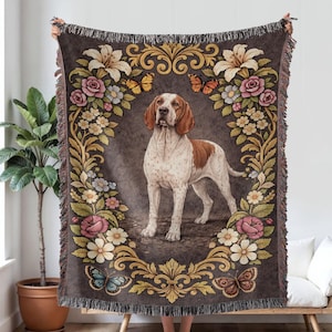May include: A woven throw blanket featuring a brown and white dog in a floral frame with butterflies. The blanket has a dark brown background with a fringed edge. The dog is standing in the centre of the blanket.