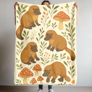 May include: A cream-colored blanket featuring a repeating pattern of cartoon platypuses, mushrooms, and foliage in shades of brown, orange, and green. The blanket has a soft, plush texture, perfect for snuggling.