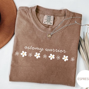 May include: Espresso-colored t-shirt with the words "ostomy warrior" in white script, accompanied by a row of white daisy graphics. A silver necklace with a pendant rests on the shirt. The shirt is a Comfort Colors brand.