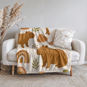 Puede incluir: Una manta beige con un patr&oacute;n repetido de carpinchos marrones, arco&iacute;ris y formas abstractas. La manta est&aacute; sobre un sof&aacute; blanco, con un coj&iacute;n decorativo. El dise&ntilde;o tiene un tema caprichoso inspirado en la naturaleza.