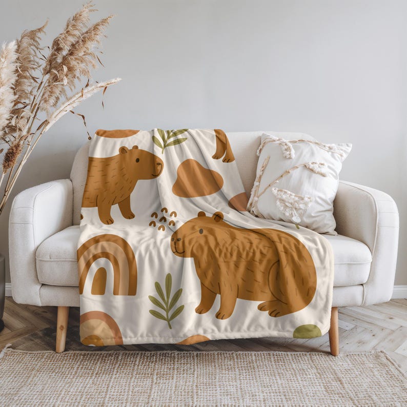 Puede incluir: Una manta beige con un patr&oacute;n repetido de carpinchos marrones, arco&iacute;ris y formas abstractas. La manta est&aacute; sobre un sof&aacute; blanco, con un coj&iacute;n decorativo. El dise&ntilde;o tiene un tema caprichoso inspirado en la naturaleza.