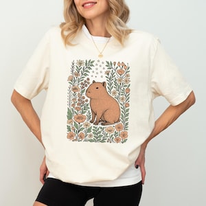 Puede incluir: Camiseta color crema con una ilustraci&oacute;n de carpincho marr&oacute;n rodeada de flores y follaje en tonos melocot&oacute;n, verde y crema. El carpincho est&aacute; sentado. La camiseta tiene mangas cortas.