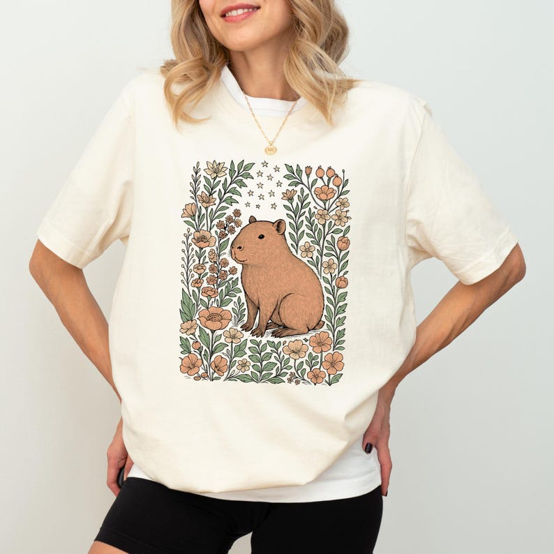 Puede incluir: Camiseta color crema con una ilustraci&oacute;n de carpincho marr&oacute;n rodeada de flores y follaje en tonos melocot&oacute;n, verde y crema. El carpincho est&aacute; sentado. La camiseta tiene mangas cortas.