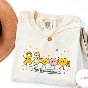 Puede incluir: Una camiseta de color marfil con un gráfico de un lazo de concienciación amarillo, una columna vertebral, una vejiga, un cerebro y un corazón, todos con caras sonrientes y tomados de la mano. Debajo, una pancarta muestra "SPINA BIFIDA AWARENESS".