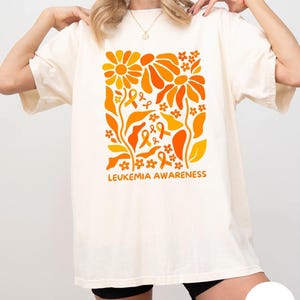 May include: Ivory-colored t-shirt featuring a floral design in shades of orange and yellow, with the words "LEUKEMIA AWARENESS" printed below. The design includes flowers, ribbons, and leaves. The shirt is a casual, oversized fit.