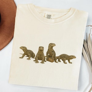 Puede incluir: Camiseta color crema con un gráfico de cuatro lagartos en diferentes poses. Los lagartos están representados en tonos marrones y beige, con escamas y detalles. La camiseta tiene cuello redondo y mangas cortas.