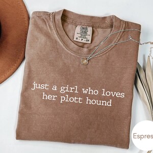 May include: Espresso-coloured t-shirt with the text "just a girl who loves her plott hound" in white. A silver necklace with a pendant rests on the shirt. The shirt is folded and displayed on a white surface.