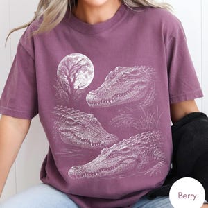 May include: A berry-colored short-sleeve t-shirt featuring a white graphic print of three alligators positioned in a swampy setting under a large full moon and a silhouetted tree. The design displays detailed textures of the alligator scales.