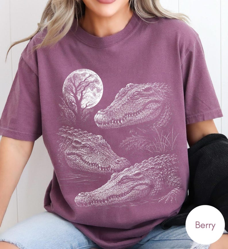 May include: A berry-colored short-sleeve t-shirt featuring a white graphic print of three alligators positioned in a swampy setting under a large full moon and a silhouetted tree. The design displays detailed textures of the alligator scales.