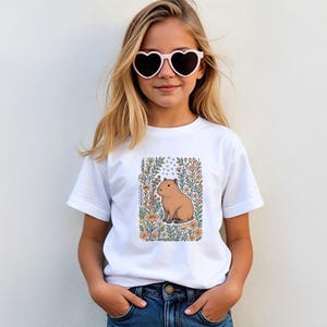 Puede incluir: Camiseta blanca con una ilustraci&oacute;n de carpincho rodeado de flores y follaje. El carpincho es marr&oacute;n y las flores son naranjas y amarillas. La persona lleva gafas de sol en forma de coraz&oacute;n.