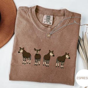 Op de afbeelding: Een bruin t-shirt met korte mouwen en een ronde hals, voorzien van een afbeelding van vier okapi's op een rij. Het shirt heeft een Comfort Colors merklabel in de binnenkraag met de tekst S P CH. Een zilveren ketting ligt op de stof.