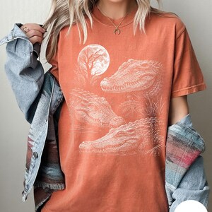 May include: A rust-colored short-sleeve t-shirt featuring a white graphic print of three alligators and a full moon. The shirt is worn with a light blue denim jacket that has a multi-colored patterned trim along the sleeve and hemline.