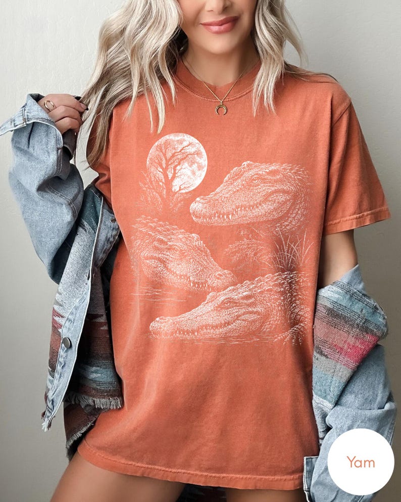 May include: A rust-colored short-sleeve t-shirt featuring a white graphic print of three alligators and a full moon. The shirt is worn with a light blue denim jacket that has a multi-colored patterned trim along the sleeve and hemline.