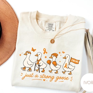 Può includere: T-shirt color avorio con un cartone animato di quattro anatre che camminano in fila, ognuna con accessori e nastri arancioni. Il testo "just a strong goose" è stampato sotto. Una collana d'oro è drappeggiata sulla maglietta.