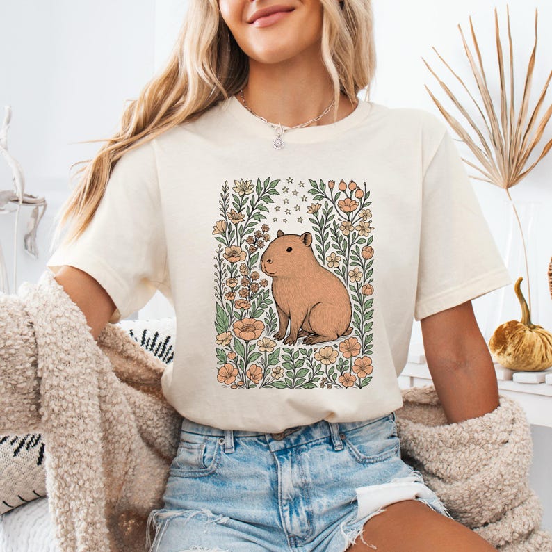 Puede incluir: Camiseta color crema con una ilustraci&oacute;n de carpincho marr&oacute;n rodeada de flores y hojas en un marco rectangular. El dise&ntilde;o incluye elementos florales melocot&oacute;n y verdes y peque&ntilde;as estrellas. La camiseta la lleva una persona.