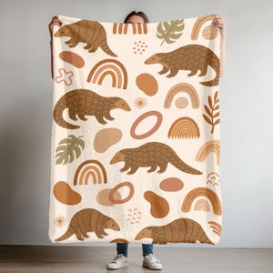 May include: A cream-colored blanket featuring a repeating pattern of brown pangolins, rainbows, and abstract shapes. The blanket has a soft, plush texture and is designed for comfort and warmth. The design incorporates earth tones and a whimsical aesthetic.