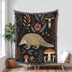 May include: A woven throw blanket featuring a pangolin illustration surrounded by mushrooms and foliage in shades of brown, beige, and teal on a black background. The blanket has a fringed edge and is held up by a person.